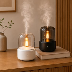 May include: Two contemporary essential oil diffusers, one white and one black, each with a clear glass dome. The diffusers have a warm, glowing light and emit vapor. They are placed on a wooden surface, suggesting a home decor theme. The units are likely to be measured in centimetres.