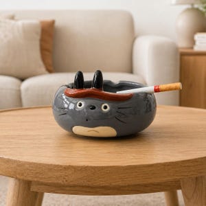 Peut inclure: Cendrier en céramique grise en forme de personnage de dessin animé, avec des oreilles noires, un bandeau rouge et des yeux blancs. Une cigarette allumée repose dans le cendrier. Le cendrier est sur une table ronde en bois.