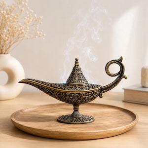 Op de afbeelding: Een antieke olielamp van messing met ingewikkelde bloemengravures, een puntig deksel en een gebogen handvat. Witte rookpluimen stijgen op uit de bovenkant van de lamp, die op een ronde voet met gedetailleerde metalen patronen staat.
