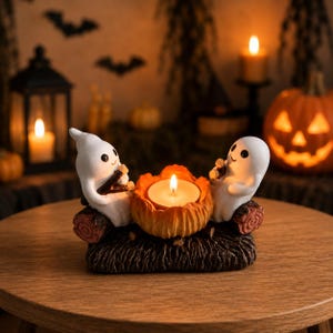 Può includere: Un portacandele decorativo con due statuette di fantasmi bianchi sedute su una base a forma di tronco marrone scuro. I fantasmi sono posizionati ai lati di un supporto centrale a forma di zucca arancione con una candelina accesa.
