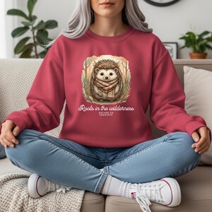 Peut inclure: Un sweat-shirt bordeaux avec une illustration détaillée d'un hérisson portant une écharpe, dans une scène de forêt. Le texte "Roots in the wilderness, Escape to Serenity" est imprimé en dessous. Le sweat-shirt est de couleur bordeaux unie.