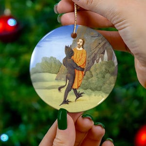 Puede incluir: Adorno navideño redondo de cerámica con una pintura de estilo renacentista de dos figuras. Una figura viste de naranja, la otra es un demonio negro. El adorno tiene una cuerda dorada para colgar y un fondo borroso de árbol de Navidad.