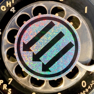 May include: Close-up of a black rotary dial telephone. The dial features white lettering for numbers and letters. A circular sticker with a holographic, glittery background and three black arrows is centered on the dial.