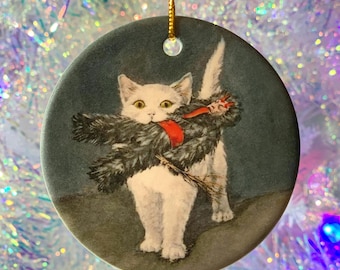 Peut inclure: Une boule de Noël en céramique blanche représentant un chat blanc tenant un Père Noël en tissu noir et rouge dans sa gueule. Le chat est debout sur un fond bleu.