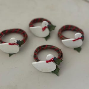 May include: Four decorative napkin rings. Each ring features a white wooden duck with a red scarf and a green leaf accent. The rings are set on a white surface. The rings are made of a brown woven material with red accents.