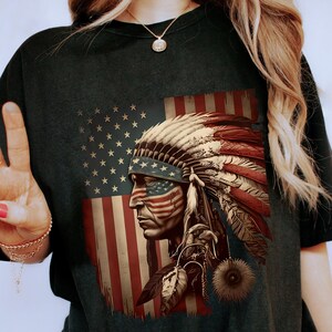 May include: Dark green t-shirt featuring a Native American chief with a headdress and American flag design. The graphic is in shades of red, white, and blue. The shirt is a relaxed fit.