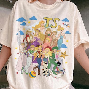 May include: A cream-colored short-sleeve t-shirt featuring a large graphic print of various animated characters dressed in costumes. The design includes the letters TS, a rocket, a yellow ball, and a sheriff star, all set against a background of blue clouds.