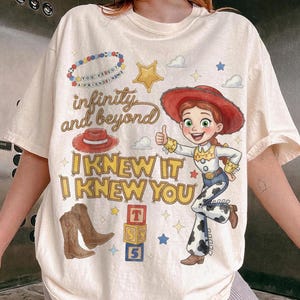 May include: A cream-colored t-shirt featuring a graphic of a cowgirl character, a red hat, brown cowboy boots, and alphabet blocks. The text reads infinity and beyond, I KNEW IT I KNEW YOU, and YOU'VE GOT A FRIEND IN ME on a beaded bracelet graphic.