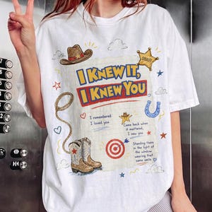 May include: A white t-shirt featuring a graphic print with the text I KNEW IT, I KNEW YOU, along with illustrations of a cowboy hat, a sheriff star, a lasso, a horseshoe, a target, and cowboy boots, plus additional handwritten-style text.