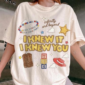 May include: A cream-colored short-sleeve t-shirt featuring a graphic print with the text "I KNEW IT I KNEW YOU" in yellow, "infinity and beyond" in black, and "YOU'VE GOT A FRIEND IN ME" on a beaded bracelet illustration, plus cowboy boots, a hat, and blocks.