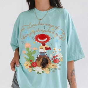 May include: A light blue t-shirt featuring a graphic of a cowgirl in a red hat riding a horse, surrounded by cacti and colorful flowers. The text "Love has ways of bringing things back to life" is printed in a cursive font above the illustration.