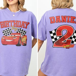 Op de afbeelding: Een lichtpaars T-shirt met een afbeelding van een rode raceauto met de tekst "BIRTHDAY BOY" en geblokte vlaggen. De achterkant van de shirt toont de naam "DANIEL" boven een groot nummer "2" met een geblokt vlagontwerp.