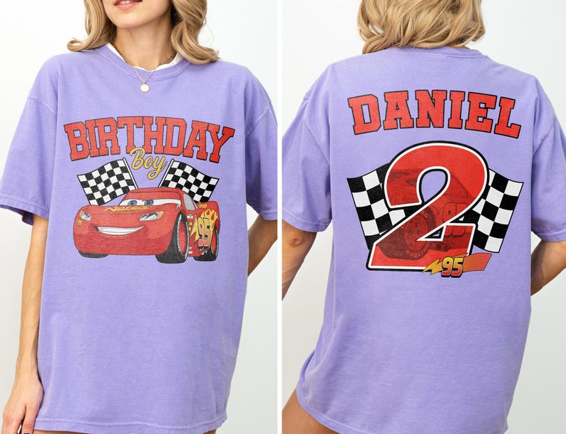 Op de afbeelding: Een lichtpaars T-shirt met een afbeelding van een rode raceauto met de tekst "BIRTHDAY BOY" en geblokte vlaggen. De achterkant van de shirt toont de naam "DANIEL" boven een groot nummer "2" met een geblokt vlagontwerp.