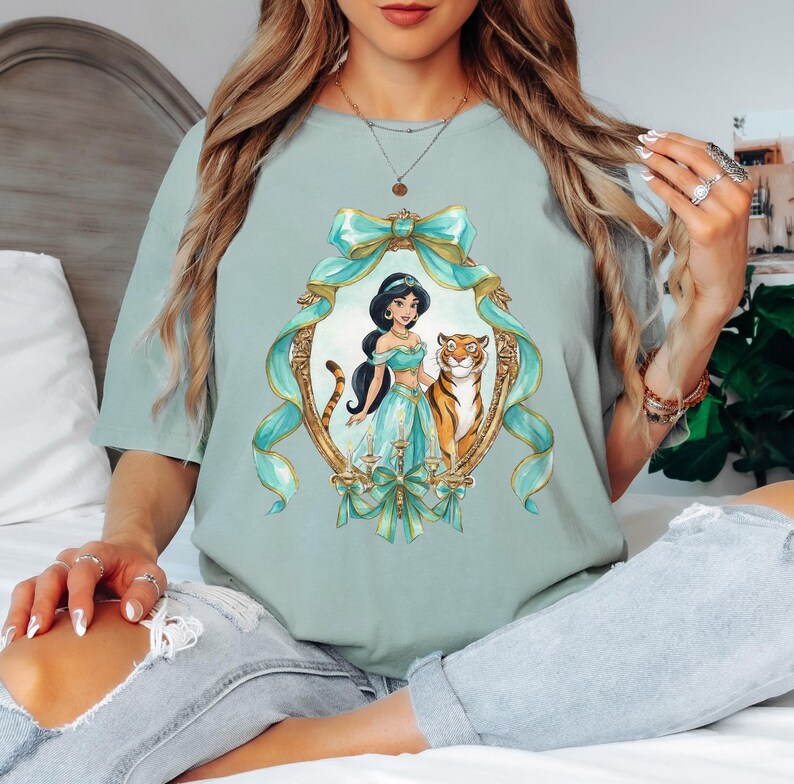 May include: A light green t-shirt featuring a watercolor illustration of a princess and a tiger inside a gold frame with teal ribbons and bows. The princess wears a teal outfit and has black hair. The tiger is orange and white.