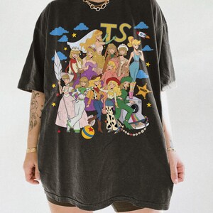 May include: A charcoal gray oversized t-shirt featuring a large graphic print of various animated characters in diverse outfits, including a sheriff star, a camera, and a colorful ball. The letters TS are printed in yellow at the top center.