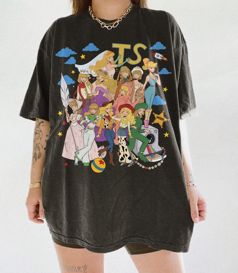 May include: A charcoal gray oversized t-shirt featuring a large graphic print of various animated characters in diverse outfits, including a sheriff star, a camera, and a colorful ball. The letters TS are printed in yellow at the top center.
