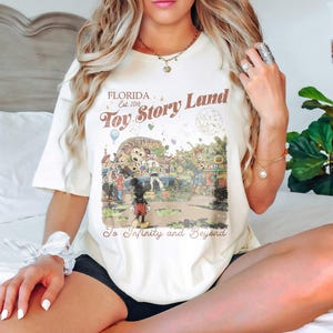 May include: Cream-colored t-shirt featuring a vintage-style graphic of "Toy Story Land" in Florida, with Mickey Mouse and other characters. The text "To Infinity and Beyond" is also included.