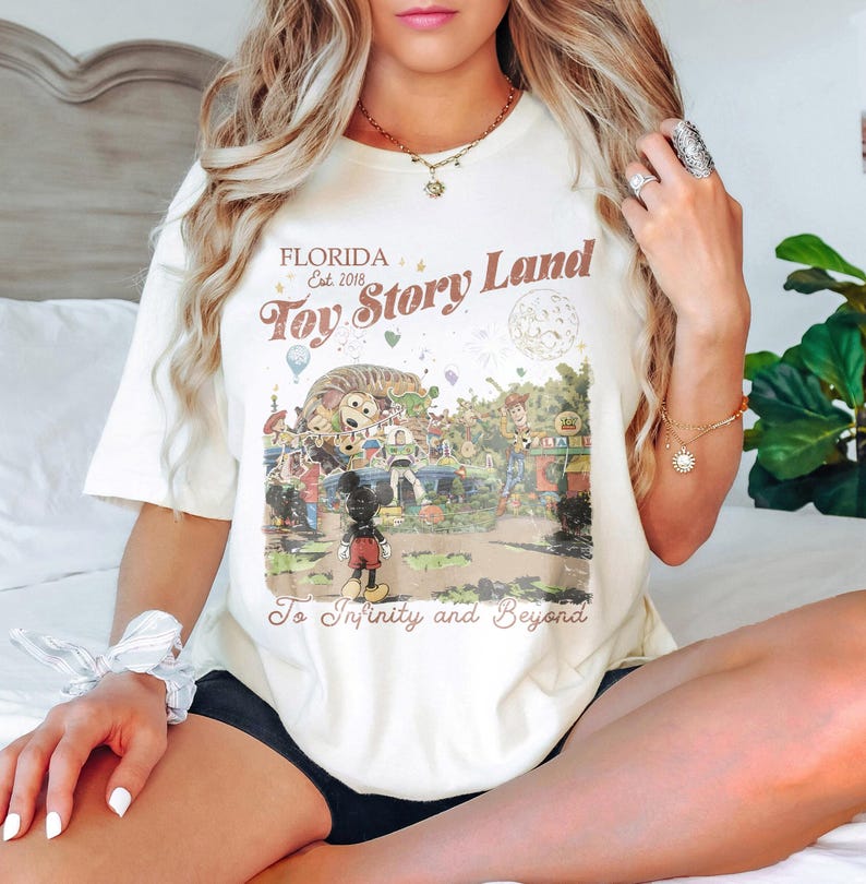 May include: Cream-colored t-shirt featuring a vintage-style graphic of "Toy Story Land" in Florida, with Mickey Mouse and other characters. The text "To Infinity and Beyond" is also included.