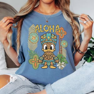 May include: Blue t-shirt with the word "ALOHA" in yellow block letters. The shirt features a cartoon duck wearing a tribal outfit, surrounded by tropical-themed graphics in shades of green, orange, and yellow.