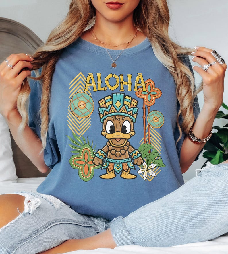 May include: Blue t-shirt with the word "ALOHA" in yellow block letters. The shirt features a cartoon duck wearing a tribal outfit, surrounded by tropical-themed graphics in shades of green, orange, and yellow.