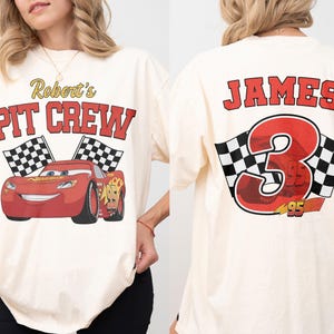 Op de afbeelding: Cr&egrave;mekleurig T-shirt met een rode raceauto-afbeelding en de tekst "Robert's PIT CREW" op de voorkant. De achterkant van het shirt toont de naam "JAMES" boven een groot nummer "3" en geblokte vlaggen.