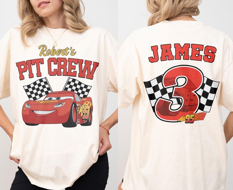Op de afbeelding: Cr&egrave;mekleurig T-shirt met een rode raceauto-afbeelding en de tekst "Robert's PIT CREW" op de voorkant. De achterkant van het shirt toont de naam "JAMES" boven een groot nummer "3" en geblokte vlaggen.