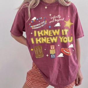 May include: A maroon short-sleeve t-shirt featuring a graphic print with the text "I KNEW IT I KNEW YOU", "infinity and beyond", and "YOU'VE GOT A FRIEND IN ME". The design includes illustrations of cowboy boots, a hat, blocks, and stars.