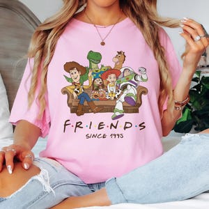 May include: Pink t-shirt featuring a graphic of characters from the animated movie Toy Story, seated on a brown couch. The word "FRIENDS" is printed below the characters, with "SINCE 1995" underneath.