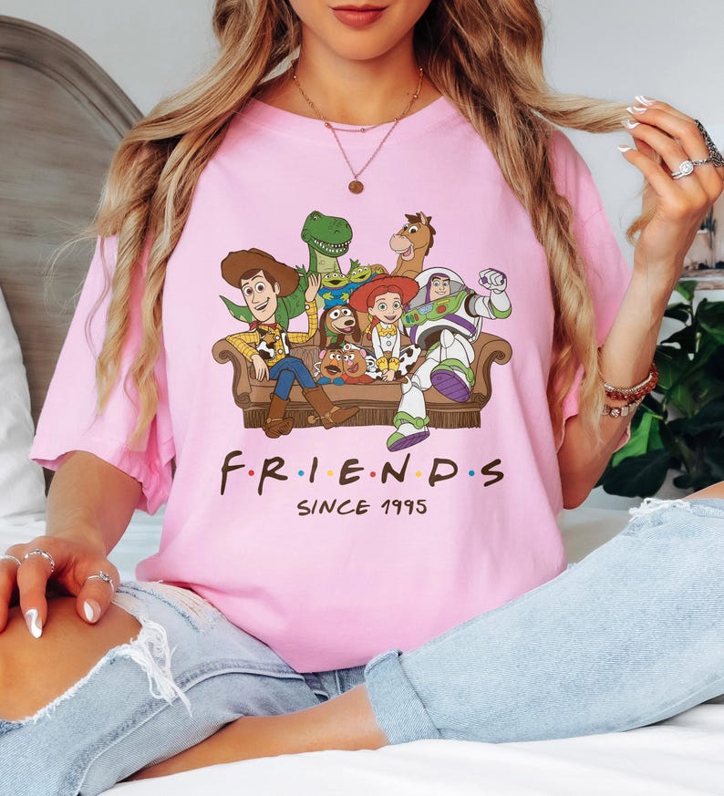 May include: Pink t-shirt featuring a graphic of characters from the animated movie Toy Story, seated on a brown couch. The word "FRIENDS" is printed below the characters, with "SINCE 1995" underneath.