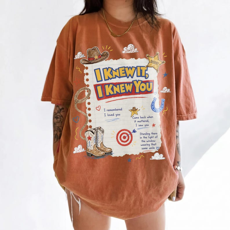 May include: An oversized burnt orange t-shirt featuring a graphic print of a notebook page with the text I KNEW IT, I KNEW YOU. The design includes illustrations of a cowboy hat, a sheriff badge, a lasso, a horseshoe, boots, and a target.