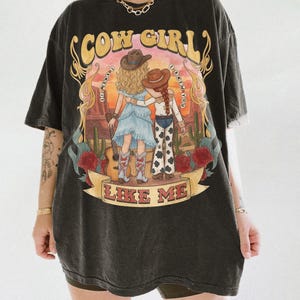 May include: A black short-sleeve t-shirt featuring a graphic print of two figures in cowboy hats standing in a desert landscape. The text reads COW GIRL at the top, I KNEW YOU on both sides, and LIKE ME on a banner at the bottom with red roses.