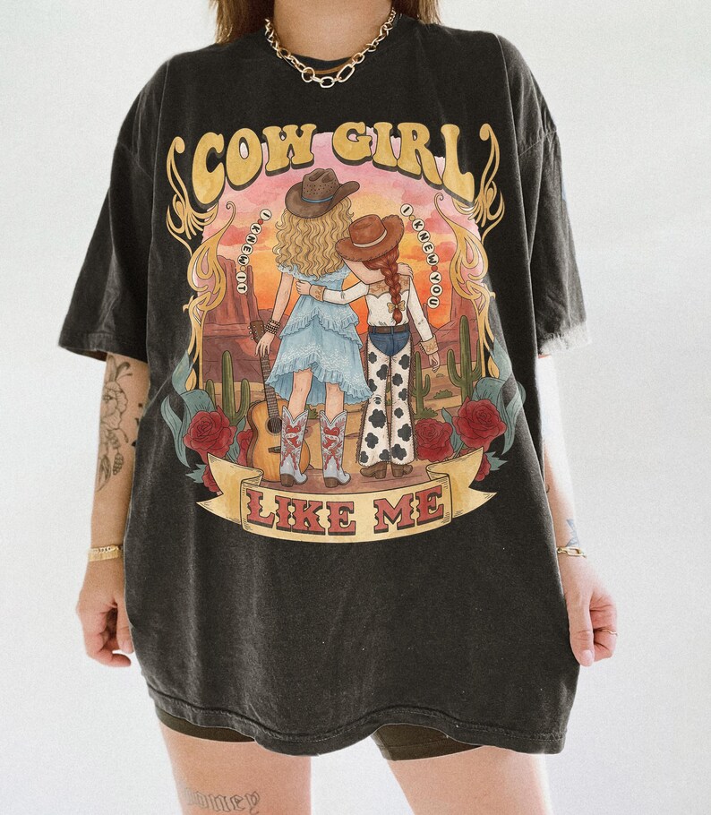 May include: A black short-sleeve t-shirt featuring a graphic print of two figures in cowboy hats standing in a desert landscape. The text reads COW GIRL at the top, I KNEW YOU on both sides, and LIKE ME on a banner at the bottom with red roses.
