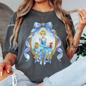 May include: Dark gray t-shirt featuring a framed illustration of Cinderella in a blue gown with two mice. The frame is adorned with a blue bow and ribbons. The shirt is a comfortable, casual style.