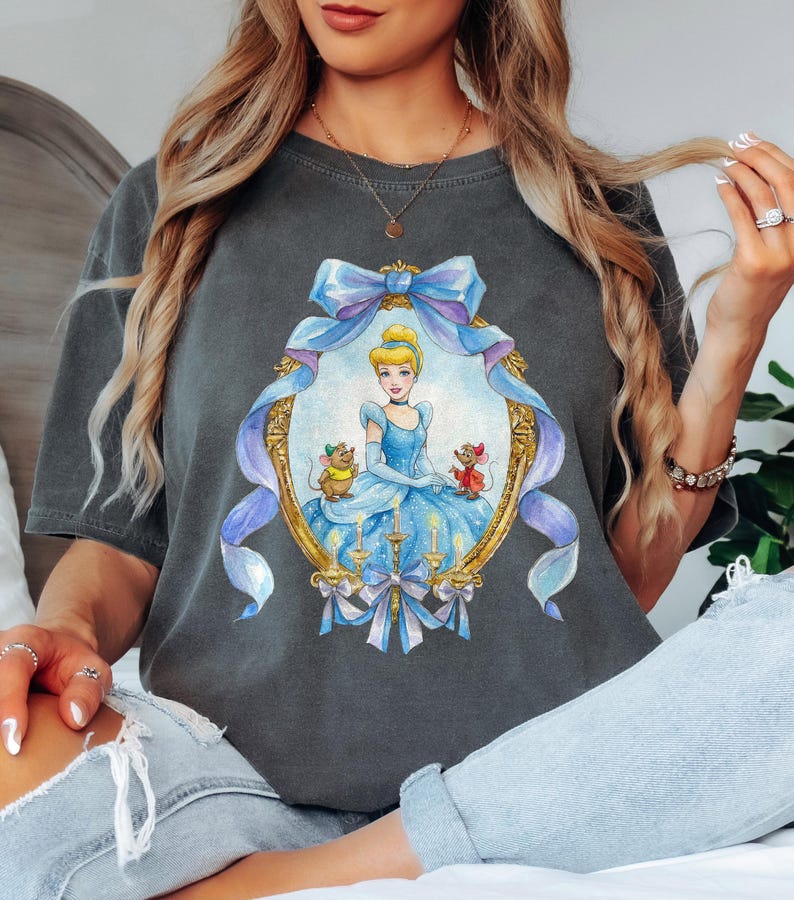 May include: Dark gray t-shirt featuring a framed illustration of Cinderella in a blue gown with two mice. The frame is adorned with a blue bow and ribbons. The shirt is a comfortable, casual style.
