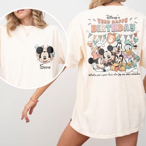 Puede incluir: Camiseta color crema con un gr&aacute;fico de Mickey Mouse en la parte delantera y el nombre "Steve". La parte trasera de la camiseta muestra un colorido dise&ntilde;o de cumplea&ntilde;os de Disney con Mickey, Minnie, Donald, Goofy y Daisy. El texto dice "Very Happy Birthday" y "Wishing you a year filled with joy and good memories."
