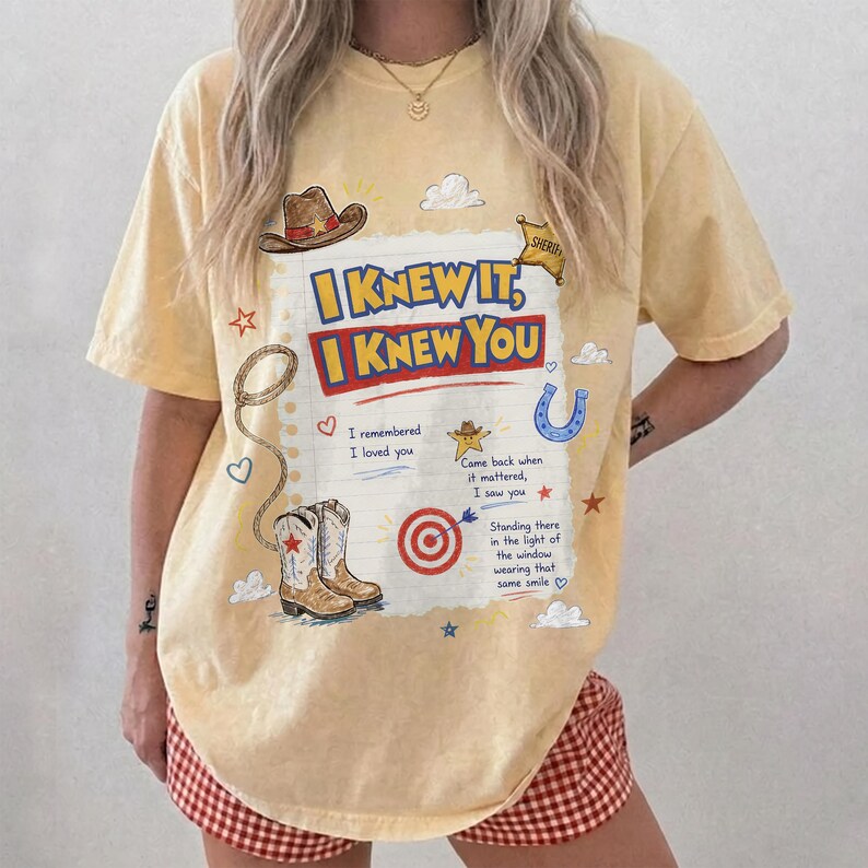 May include: A pale yellow short-sleeve t-shirt featuring a graphic print of a cowboy hat, a lasso, a pair of western boots, a target, and the text "I KNEW IT, I KNEW YOU" along with additional lyrics and small star, heart, and cloud illustrations.