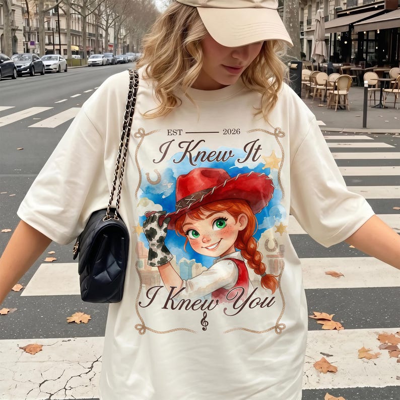 May include: An off-white t-shirt featuring a graphic of a cartoon character with red hair wearing a red cowboy hat and a cow-print glove. The text reads "EST 2026", "I Knew It", and "I Knew You" with a musical note symbol. A black quilted bag is worn.