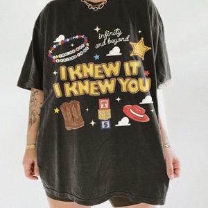 May include: A black short-sleeve t-shirt featuring a graphic print with the text "I KNEW IT I KNEW YOU", "infinity and beyond", and "YOU'VE GOT A FRIEND IN ME". The design includes illustrations of a beaded bracelet, a star, cowboy boots, blocks, and a hat.
