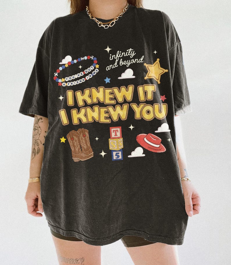 May include: A black short-sleeve t-shirt featuring a graphic print with the text "I KNEW IT I KNEW YOU", "infinity and beyond", and "YOU'VE GOT A FRIEND IN ME". The design includes illustrations of a beaded bracelet, a star, cowboy boots, blocks, and a hat.