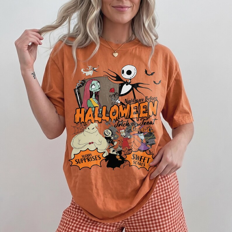 K&ouml;nnte beinhalten: Ein orangefarbenes Kurzarm-T-Shirt mit einem Grafikdruck der Charaktere aus Nightmare Before Christmas. Das Design zeigt Jack Skellington, Sally und Oogie Boogie mit dem Text Halloween, Trick or Treat und Candy Co.