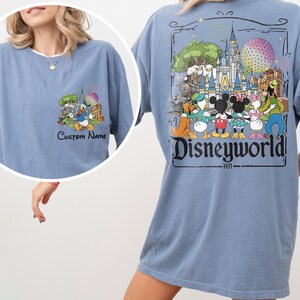 May include: A light blue t-shirt featuring a Disney World graphic. The front of the shirt has a small graphic with Donald Duck and the text "Custom Name". The back of the shirt has a larger graphic with Disney characters and the text "Disneyworld! 1971".