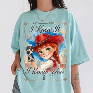 May include: A light blue t-shirt featuring a graphic of a person with red hair wearing a red cowboy hat and a cow-print glove. The text reads EST 2026, I Knew It, and I Knew You, framed by a rope border with stars and horseshoes.