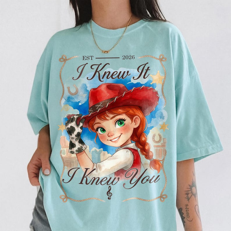 May include: A light blue t-shirt featuring a graphic of a person with red hair wearing a red cowboy hat and a cow-print glove. The text reads EST 2026, I Knew It, and I Knew You, framed by a rope border with stars and horseshoes.
