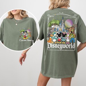 May include: Olive green t-shirt featuring a Disney World graphic on the back with Mickey Mouse and friends. The front has a smaller graphic with Mickey Mouse and the text "Custom Name".