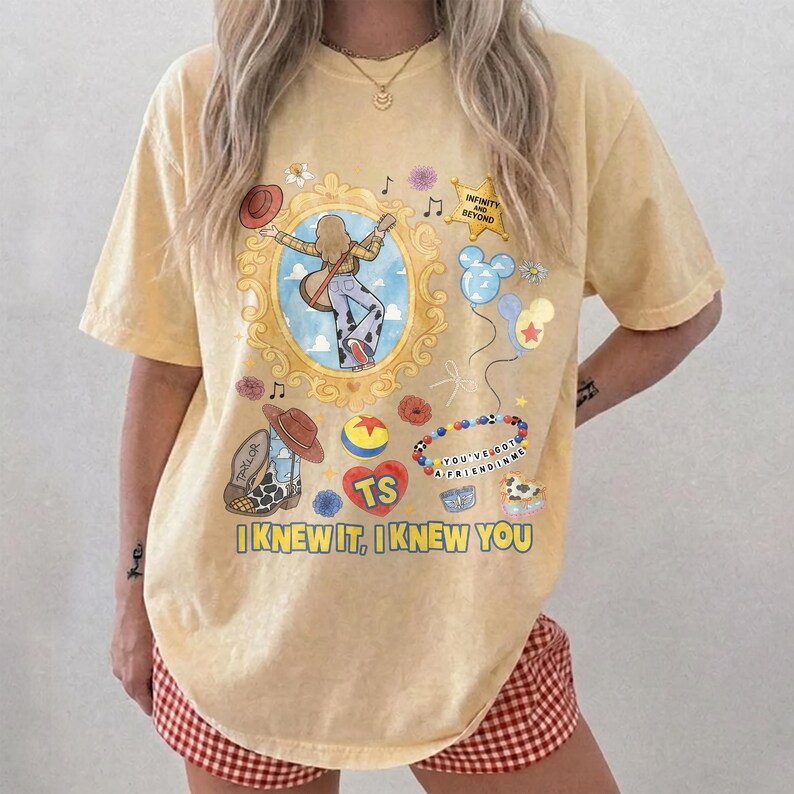 May include: A pale yellow short-sleeve t-shirt featuring a graphic print with a central gold-framed illustration of a person with a guitar, a sheriff star labeled INFINITY AND BEYOND, balloons, a beaded bracelet, and the text I KNEW IT, I KNEW YOU.