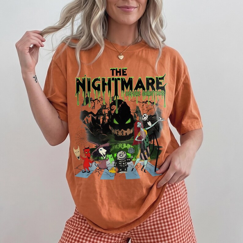 May include: Orange t-shirt featuring a graphic from "The Nightmare Before Christmas." The design includes the text "The Nightmare Before Christmas" in green, with characters like Jack Skellington and Sally. The shirt has short sleeves.