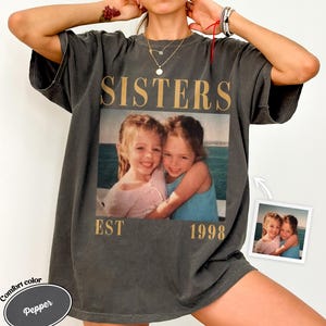 Puede incluir: Una camiseta de manga corta gris carbón con un estampado gráfico de estilo vintage de dos niñas sonriendo en la playa. El texto SISTERS está impreso en letras doradas grandes sobre la foto, con EST 1998 impreso en oro debajo de la imagen.