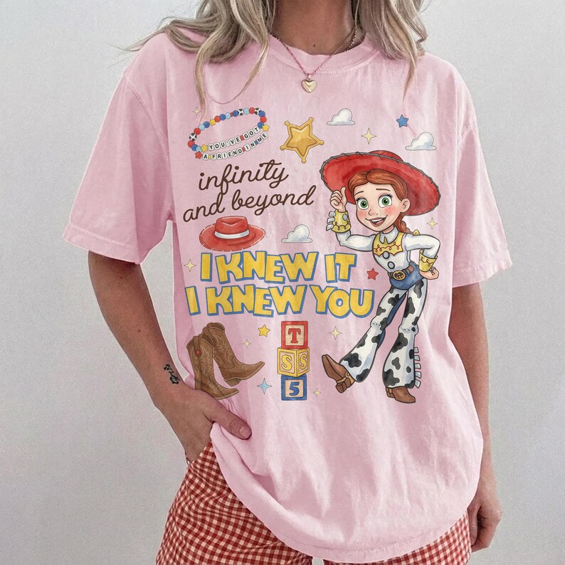Pode incluir: Uma t-shirt de manga curta rosa claro com uma impress&atilde;o gr&aacute;fica de uma personagem cowgirl, um chap&eacute;u de cowboy vermelho, botas castanhas, um colar de contas e blocos de letras. O texto diz infinity and beyond, I KNEW IT e I KNEW YOU.