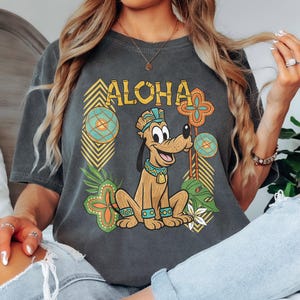 May include: Dark grey t-shirt with the word "ALOHA" and a cartoon dog in a Hawaiian theme. The dog is surrounded by tropical floral and geometric designs in green, yellow, and orange. The shirt has a relaxed fit.