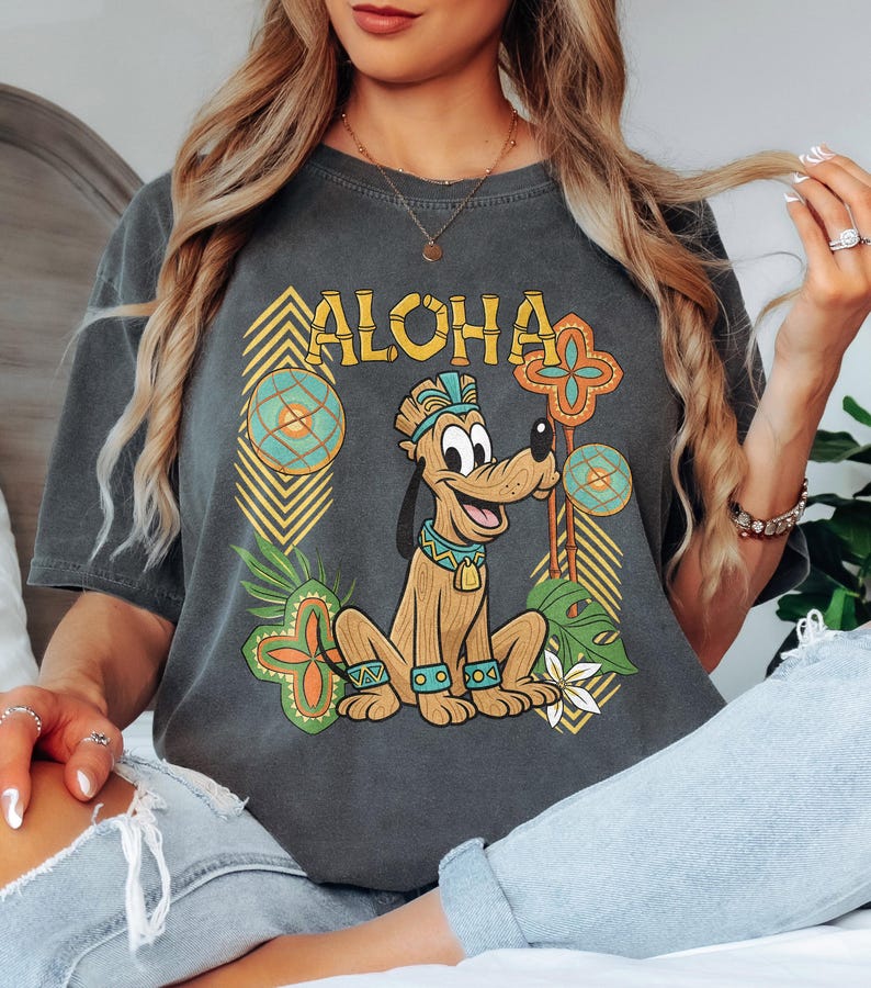 May include: Dark grey t-shirt with the word "ALOHA" and a cartoon dog in a Hawaiian theme. The dog is surrounded by tropical floral and geometric designs in green, yellow, and orange. The shirt has a relaxed fit.